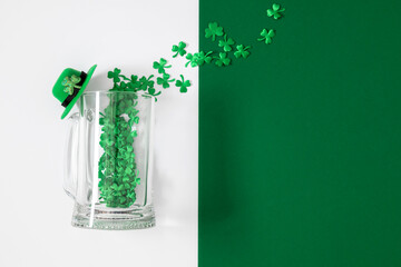St. Patrick's Day minimalist concept: Glass beer mug with green shamrock confetti and leprechaun hat on white and green background. Flat lay with copy space for Irish celebration greeting.