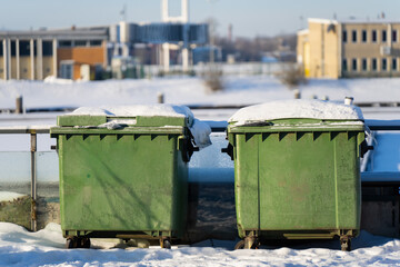 Two green wheeled trash containers covered with snow on outdoor platform, winter waste management scene with blurred industrial buildings and cold daylight background