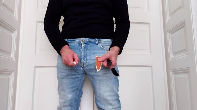 A figure in a black sweater and blue jeans stands against a white door. He holds a folding knife in one hand and its sheath in the other, demonstrating how to handle the tool safely