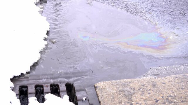 Video of an oil spill mixed with melting snow flowing into a storm drain. Iridescent surface highlights environmental pollution, water contamination, and ecological damage.