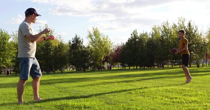 Father plays American football with son in park. Happy father throws ball to child. Active father enjoys game outdoors. Family spends time on green grass. Summer day brings fun and joy.