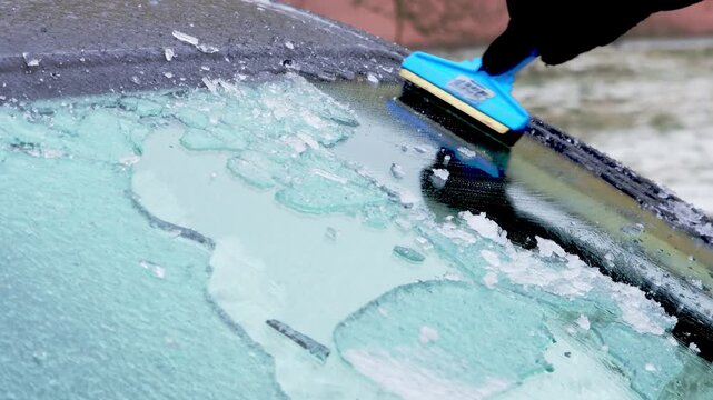 Hand using blue ice scraper to remove ice from car windshield, revealing clear glass underneath, with ice shards scattered on the surface and snow in the background