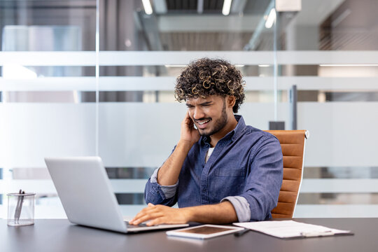 A young Indian man sits at a desk in an office and holds his hand over his ear, feeling severe pain