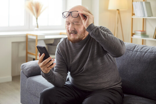 Old man with smartphone lifts eyeglasses, struggling with bad eyesight at home, navigating online internet challenges, frustration and visual difficulty in a personal domestic space.