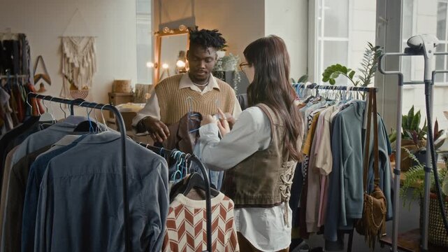 Caucaisan woman helping her African American male firend find outfit from secondhand clothes in cozy modern thrift shop