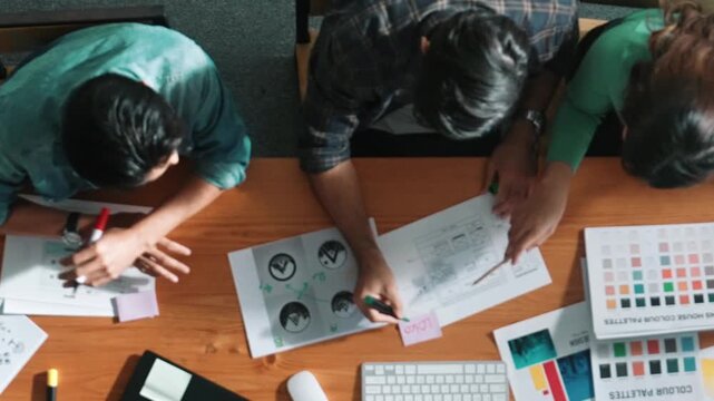 Top view of manager writing idea while team standing and giving comment. Professional designer design logo while looking at color palette and writing idea down in paper with color marker. Symposium.
