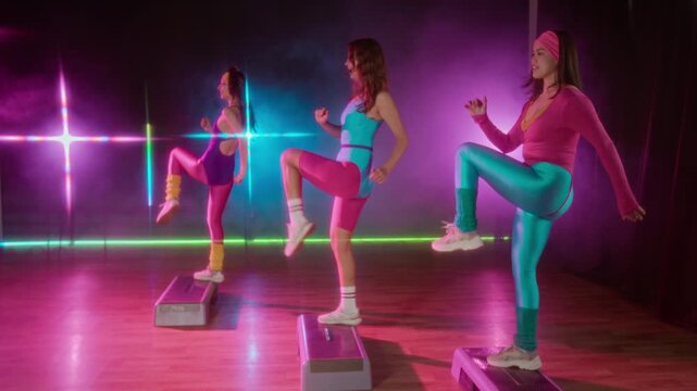 Full dolly shot of three young Caucasian women, dressed in vintage neon pink, blue and purple 1980s leggings, leotards, performing energetic step aerobics routine, with synchronized leg movements