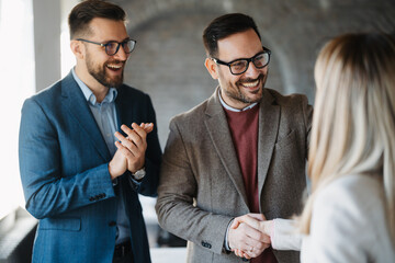 Businessman and businesswoman shaking hands in a modern office after signing a contract while a male colleague applauds in the background.

