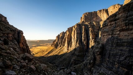 Dramatic sheer cliff face in golden hour sunlight showcasing layered geological formations and vast arid valley below
