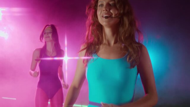 Medium shot of cheerful young Caucasian female fitness instructor, wearing microphone, leading energetic step aerobics class in gym, among neon lights and smoke, nostalgic 1980s vibe