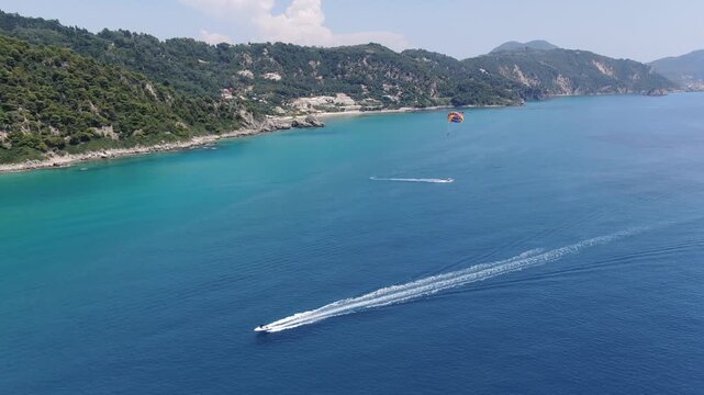 Wide aerial view of coastline with turquoise water and green hills featuring speed boat towing parasail parachute in distance under blue sky