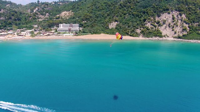Aerial view of boat towing parasailer over turquoise sea water near sandy beach and green mountains with resort hotel on coast during sunny summer day
