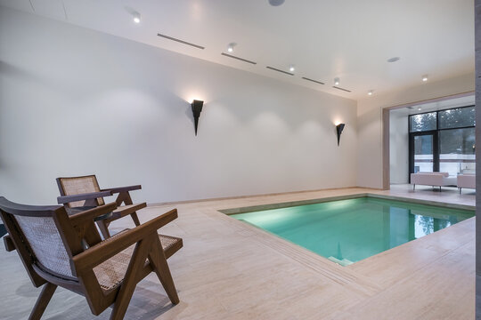A small indoor pool is surrounded by smooth walls and has wooden chairs nearby. Natural light comes in, adding to the modern ambiance of the space