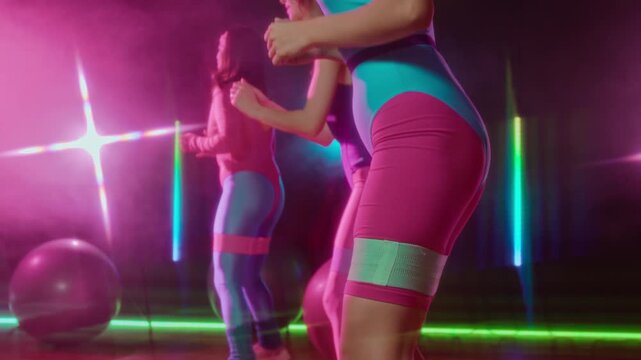 Medium shot of young women, dressed in bright blue and pink leotards and leggings, repeating rhythmic aerobics exercise with stretchy resistance bands over thighs at retro group class