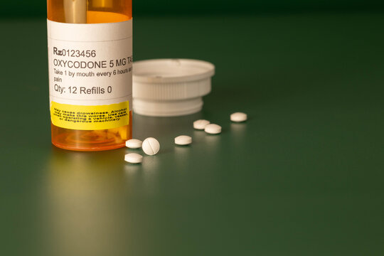Oxycodone prescription medication in yellow orange plastic bottle with round white tablets out of container on a dark green background.