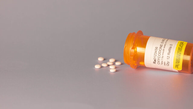 Oxycodone prescription medication in orange plastic bottle with round white tablets spilling out of container onto a gray background