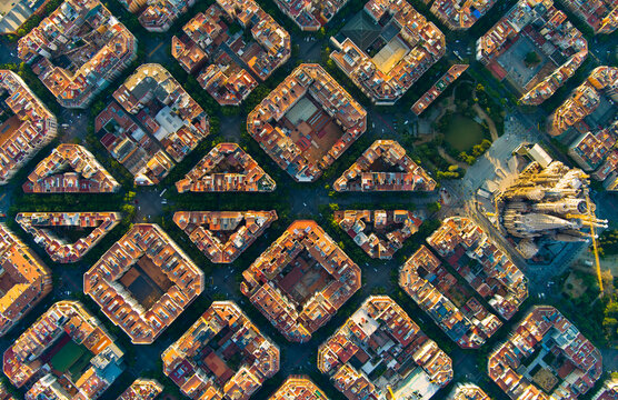 Wide aerial sunrise view of Barcelona with the Sagrada Familia visible among the Eixample grid. The city awakens under soft light, revealing its iconic layout and landmarks