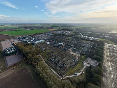 Aerial view of a sprawling National Grid Walpole Substation under a bright sky, contrasting with the muted greens and browns of the surrounding landscape, Wisbech, United Kingdom.