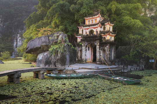 Aerial view of a pagoda nestled among verdant foliage and jagged cliffs, its ornate architecture reflected in the tranquil waters below, Ninh Hai, Ninh Binh, Vietnam.