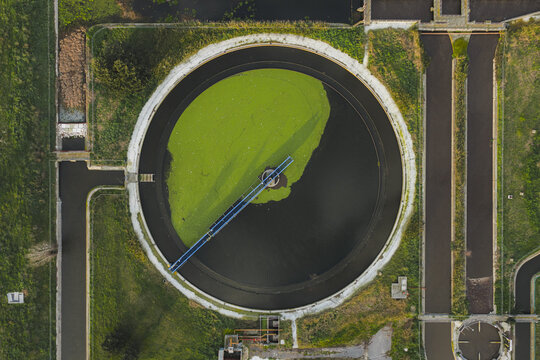 Aerial view of a circular water treatment plant with vibrant green algae contrasting against the dark water, surrounded by lush greenery, Rome, Lazio, Italy.