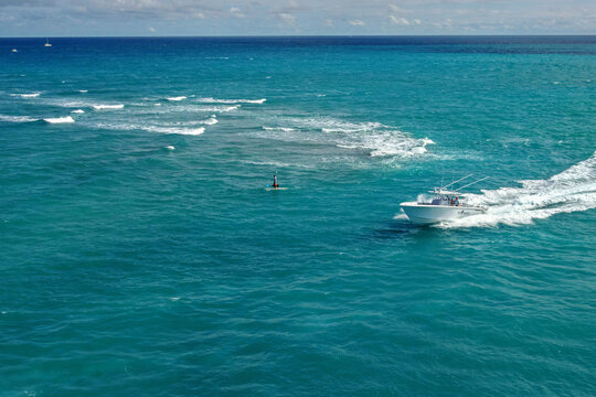 Aerial view of a boat cutting through the turquoise waters, contrasting with the white wake, near a solitary figure standing on a paddleboard, Jupiter, Florida, United States.