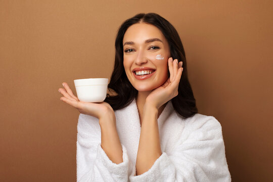 Cheerful European lady wearing white bathrobe holding jar with nourishing face cream, moisturizing skin, posing on beige studio background
