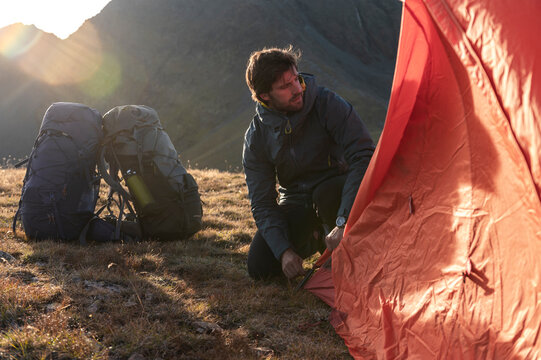 Male camper kneeling to fasten tent base in alpine terrain