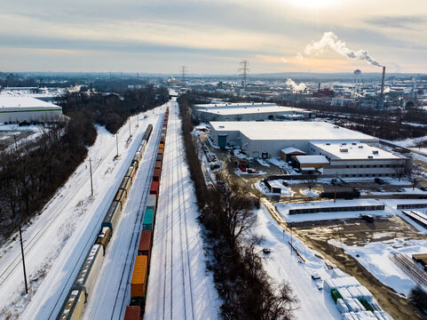 Aerial view of a long freight train cutting through a snow-laden landscape, contrasting with the industrial buildings and a smokestack puffing against the pale sky, Cincinnati, Ohio, United States.