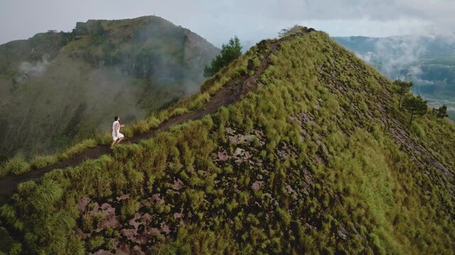 Man running along narrow ridge of Mount Batur volcano in Bali, challenging himself with endurance and fitness on trekking path during cloudy morning. Sports, recreation, healthy lifestyle. Aerial view