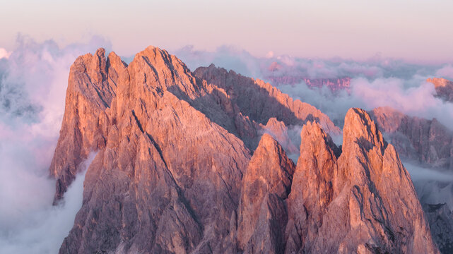 Aerial view of jagged peaks kissed by the rosy dawn, piercing through a sea of swirling clouds, Seceda, Italy.