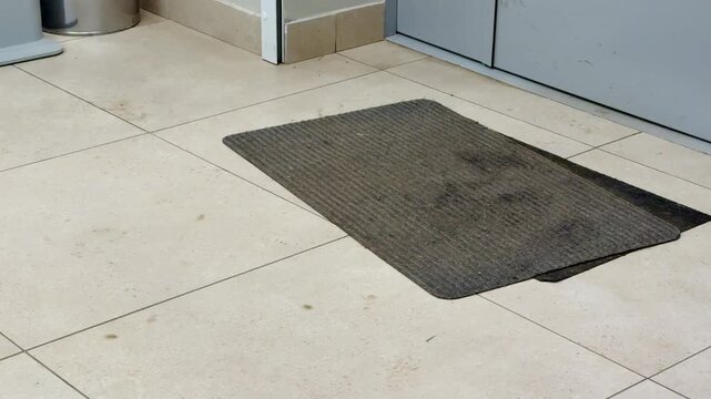Dirty gray entrance mat on tile in administrative building lobby under metal door visible stains, mud spots, worn edges, scuffed corners high foot traffic signs and dust accumulation create