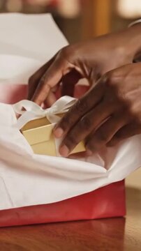 Close-up view of dark-skinned hands carefully placing a small gold gift box tied with a white ribbon into a red presentation bag lined with tissue paper.