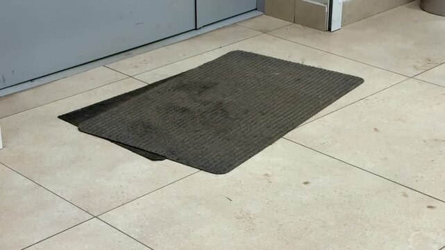 Dirty gray entrance mat on tile in administrative building lobby under metal door visible stains, mud spots, worn edges, scuffed corners high foot traffic signs and dust accumulation create
