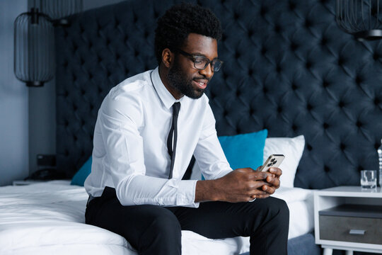 Young smiling black businessman typing smartphone sitting on bed