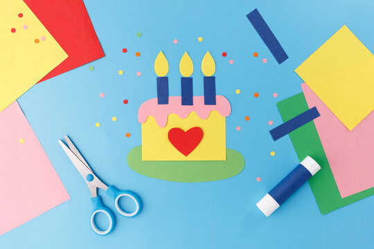 Paper craft birthday cake with candles scissors colorful sheets on blue background. Perfect for creative education, kindergarten workshops, handmade greeting card business, art supplies shop