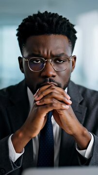 Focused African American businessman in a grey suit clasping hands in deep thought inside a modern office, illustrating corporate strategy concepts.