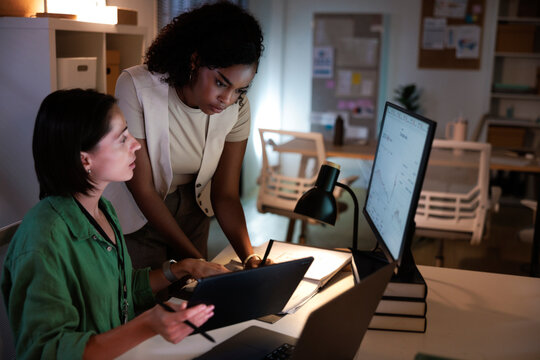 Two women collaborate on a project in a dimly lit office.