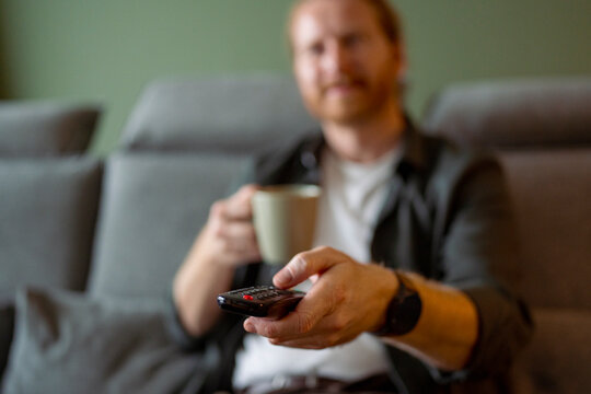 Man sits on couch and holds remote while drinking coffee
