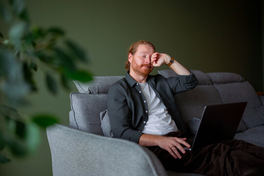 Man sitting on couch with laptop showing tired expression
