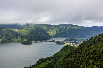 Crater lake at Sete Cidades, San Miguel Island. Lush green mountains surround the calm water. Volcanic landscape in Azores, Portugal. Cloudy weather with mist over the hills.
