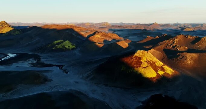 Drone view over Iceland volcanic desert at golden hour, sunlit cinder cone glowing yellow above black ash plains and mossy patches, with long shadows shaping rugged ridges.