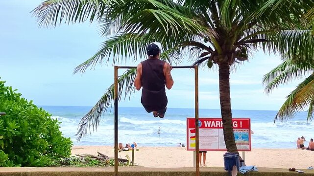 Man performing L-sit pull-ups on beach exercise bar in Phuket, Thailand. Training with the ocean as witness
