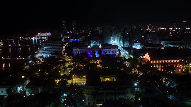 Aerial night view of the city of Recife, Pernambuco, Brazil, highlighting the illuminated urban center and the Pal&aacute;cio do Campo das Princesas along the river. 