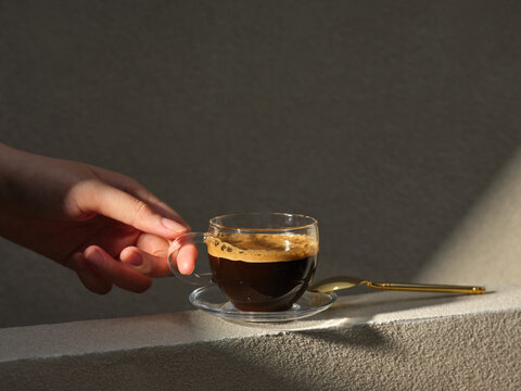 A person's hand holding a glass cup of espresso with a golden spoon