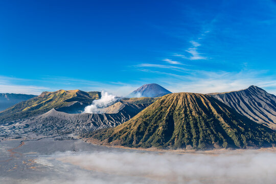 Mount Bromo, an active volcano in east Java, Indonesia as viewed from the rim of the valley. Smoke rising from the crater. Thinning fog on the sandy desert surrounding the mountains. 
