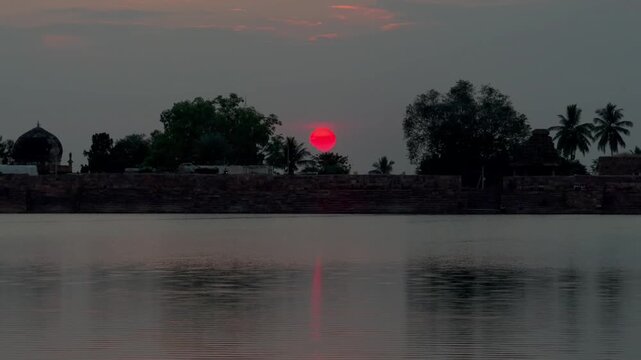 A red sunset reflecting on the agastya lake, framed by trees and a distant mosque