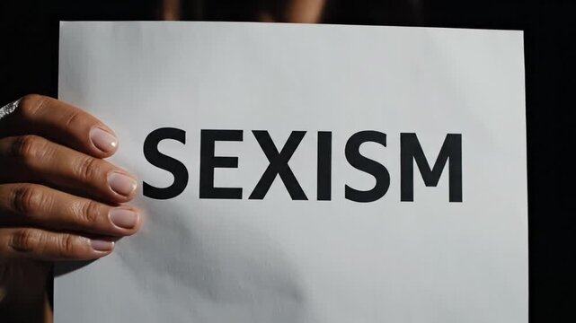 High contrast cinematic video focusing on a hand holding a white paper with bold black word SEXISM face not shown dark background dramatic soft lighting smooth slow camera movement medium close up com