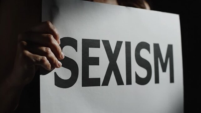 Anonymous hand holding a white protest style poster with the word SEXISM in bold black letters cinematic slow motion dark background minimal dramatic lighting soft shadows smooth camera movement mediu