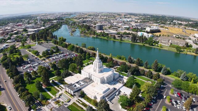 Aerial view of the Idaho Falls Temple and nearby cityscape along the Snake River, showing the white spire, surrounding parkland, trees, roads, and buildings under a clear sky.