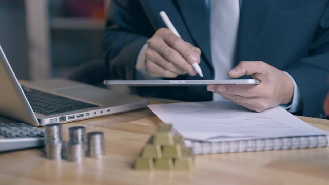 Close up of businessman using stylus pen signing digital contract on tablet with coins, gold bars, and laptop on office desk. Financial investment and online transaction concept.